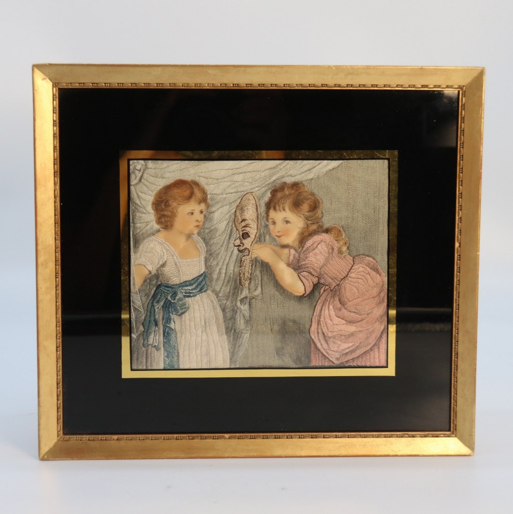 A framed Georgian silk needlework picture depicting two young children with a mask, mounted in a black eglomise frame with gold leaf detailing.