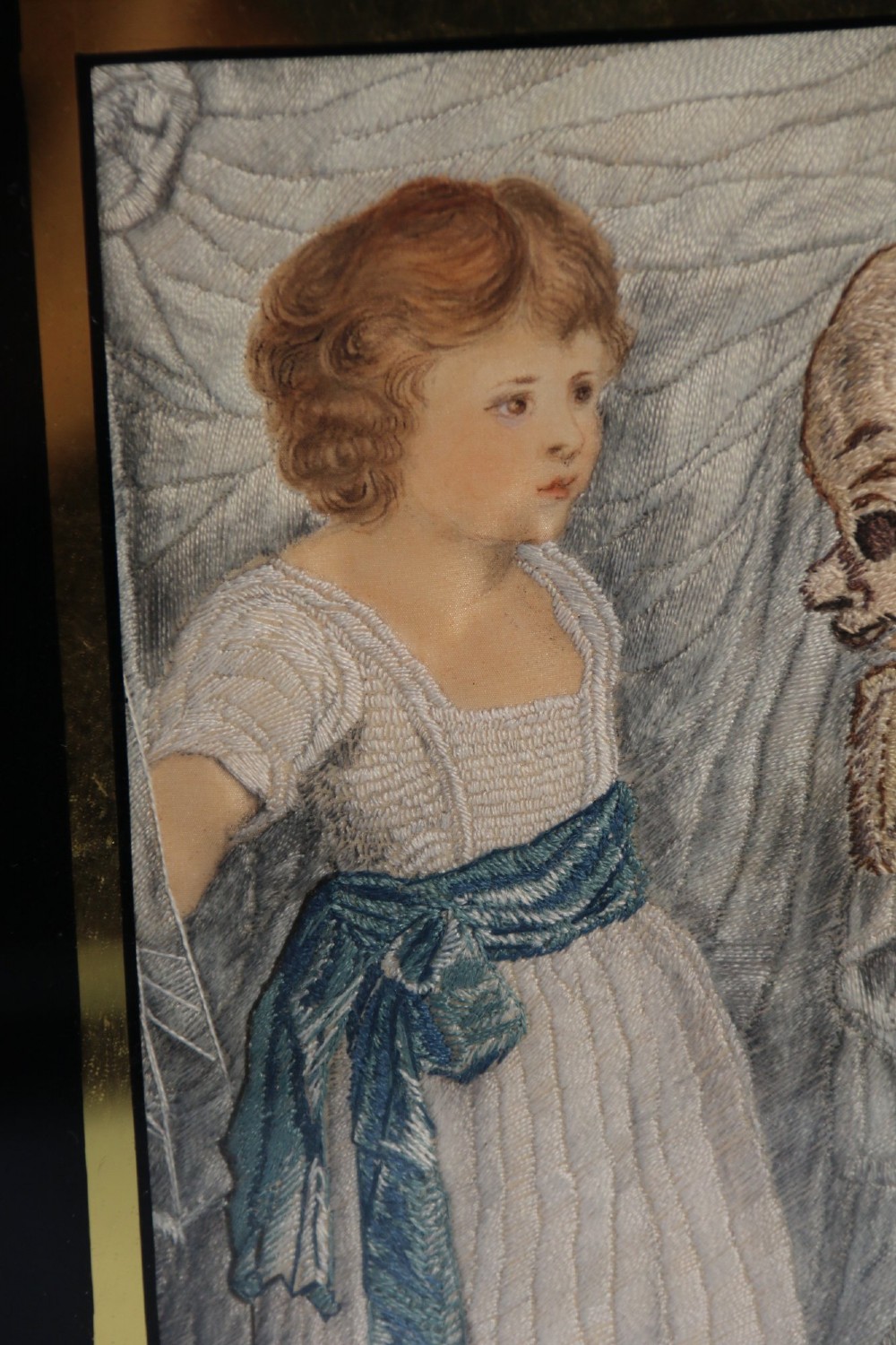 A framed Georgian silk needlework picture depicting two young children with a mask, mounted in a black eglomise frame with gold leaf detailing. View III