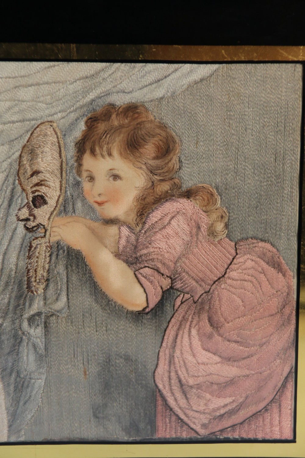 A framed Georgian silk needlework picture depicting two young children with a mask, mounted in a black eglomise frame with gold leaf detailing. View V
