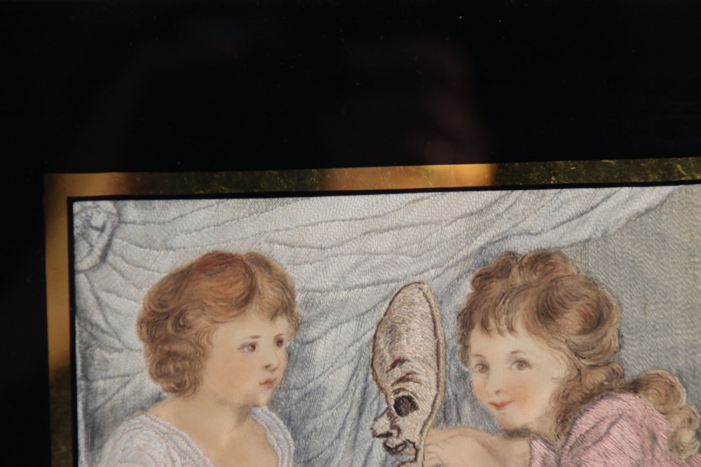 A framed Georgian silk needlework picture depicting two young children with a mask, mounted in a black eglomise frame with gold leaf detailing. View VI