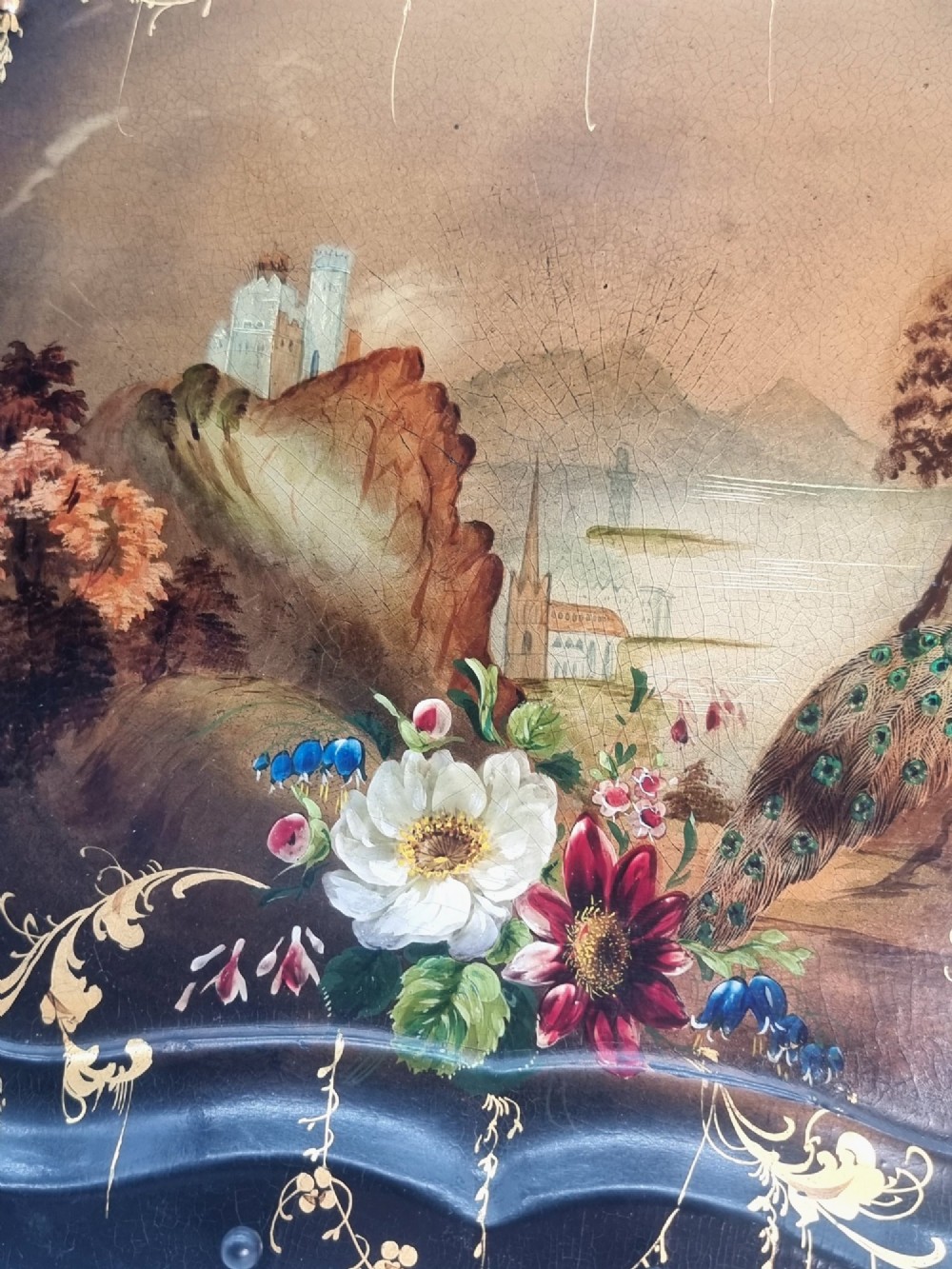 A Victorian shaped papier-mache tray featuring a hand-painted landscape with a castle and peacock surrounded by gold leaf scrollwork on a black background. View V