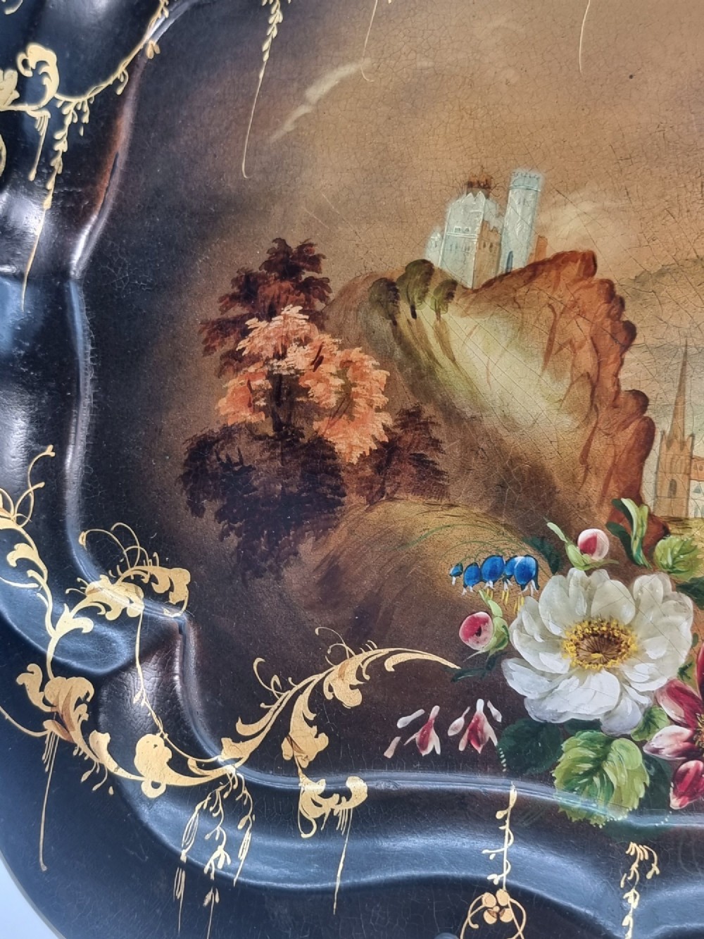 A Victorian shaped papier-mache tray featuring a hand-painted landscape with a castle and peacock surrounded by gold leaf scrollwork on a black background. View VI