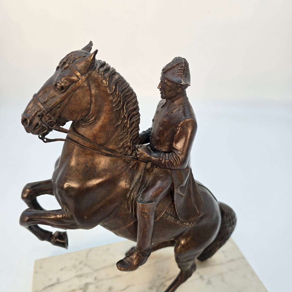 A dark bronze equestrian sculpture depicting a uniformed rider atop a rearing horse, mounted on a rectangular cream marble plinth. View X