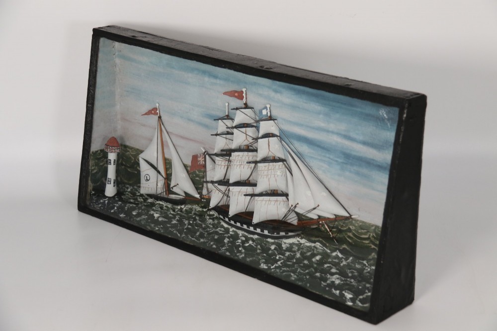 A nineteenth-century glass-fronted black wooden shadow box featuring a carved and painted diorama of two sailing ships and a lighthouse on a textured sea. View II