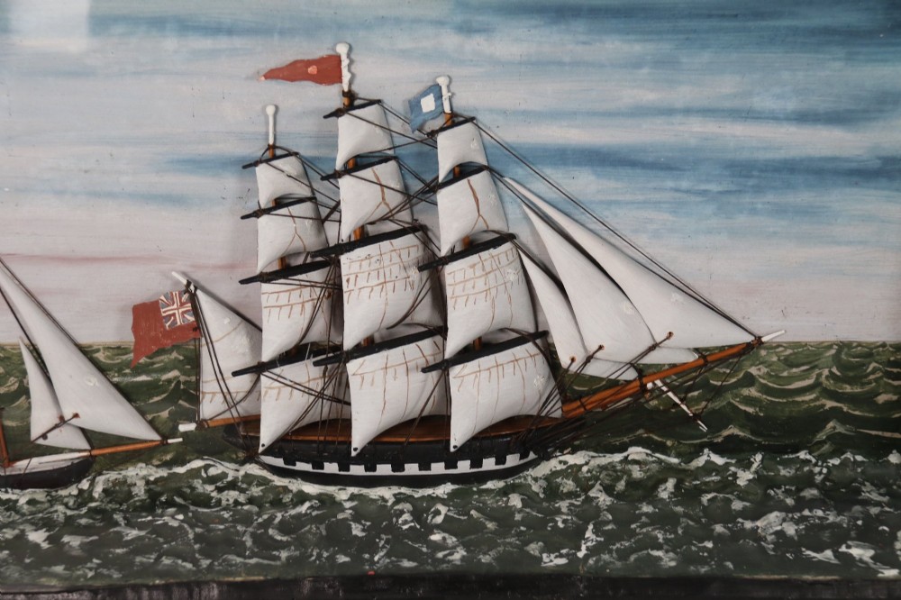 A nineteenth-century glass-fronted black wooden shadow box featuring a carved and painted diorama of two sailing ships and a lighthouse on a textured sea. View IX