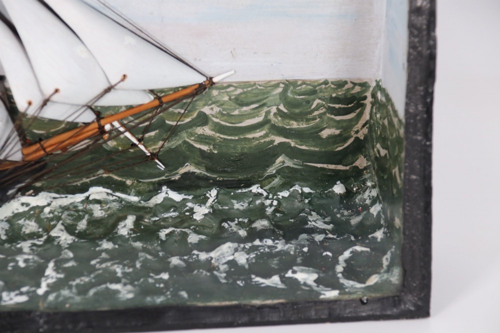 A nineteenth-century glass-fronted black wooden shadow box featuring a carved and painted diorama of two sailing ships and a lighthouse on a textured sea. View X
