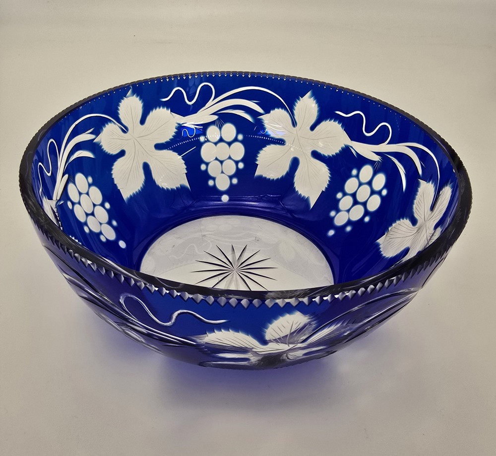 A large round Bohemian cobalt blue cut-to-clear glass bowl featuring etched grapevine leaves, grape clusters, and a serrated rim. View X