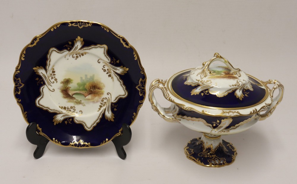 A 19th-century Mintons porcelain dessert cream tureen with lid and handles positioned alongside its matching stand, decorated with cobalt blue ground, gold gilding, and hand-painted landscape scenes.