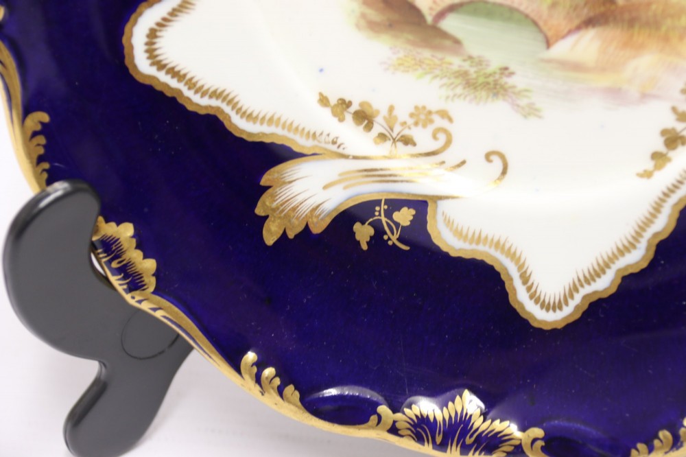 A 19th-century Mintons porcelain dessert cream tureen with lid and handles positioned alongside its matching stand, decorated with cobalt blue ground, gold gilding, and hand-painted landscape scenes. View II