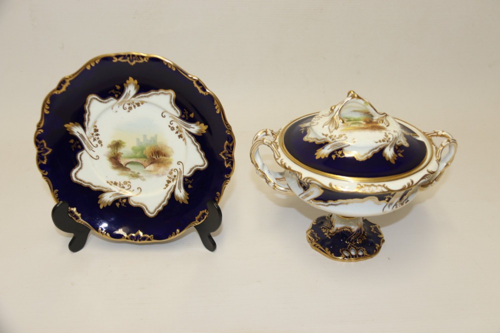 A 19th-century Mintons porcelain dessert cream tureen with lid and handles positioned alongside its matching stand, decorated with cobalt blue ground, gold gilding, and hand-painted landscape scenes. View III