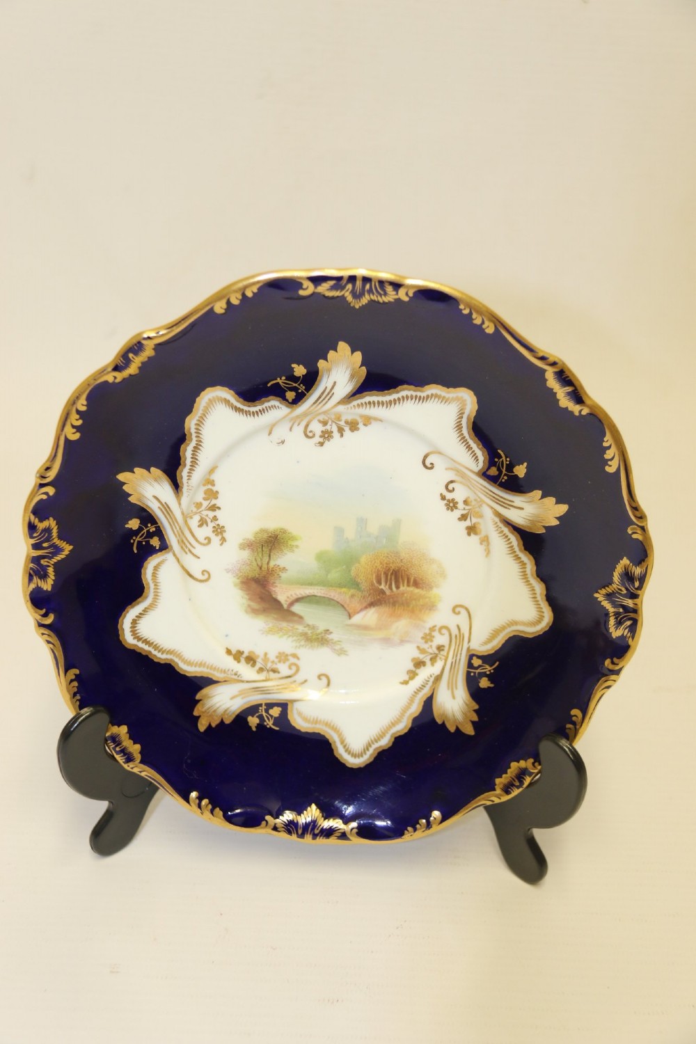 A 19th-century Mintons porcelain dessert cream tureen with lid and handles positioned alongside its matching stand, decorated with cobalt blue ground, gold gilding, and hand-painted landscape scenes. View IV