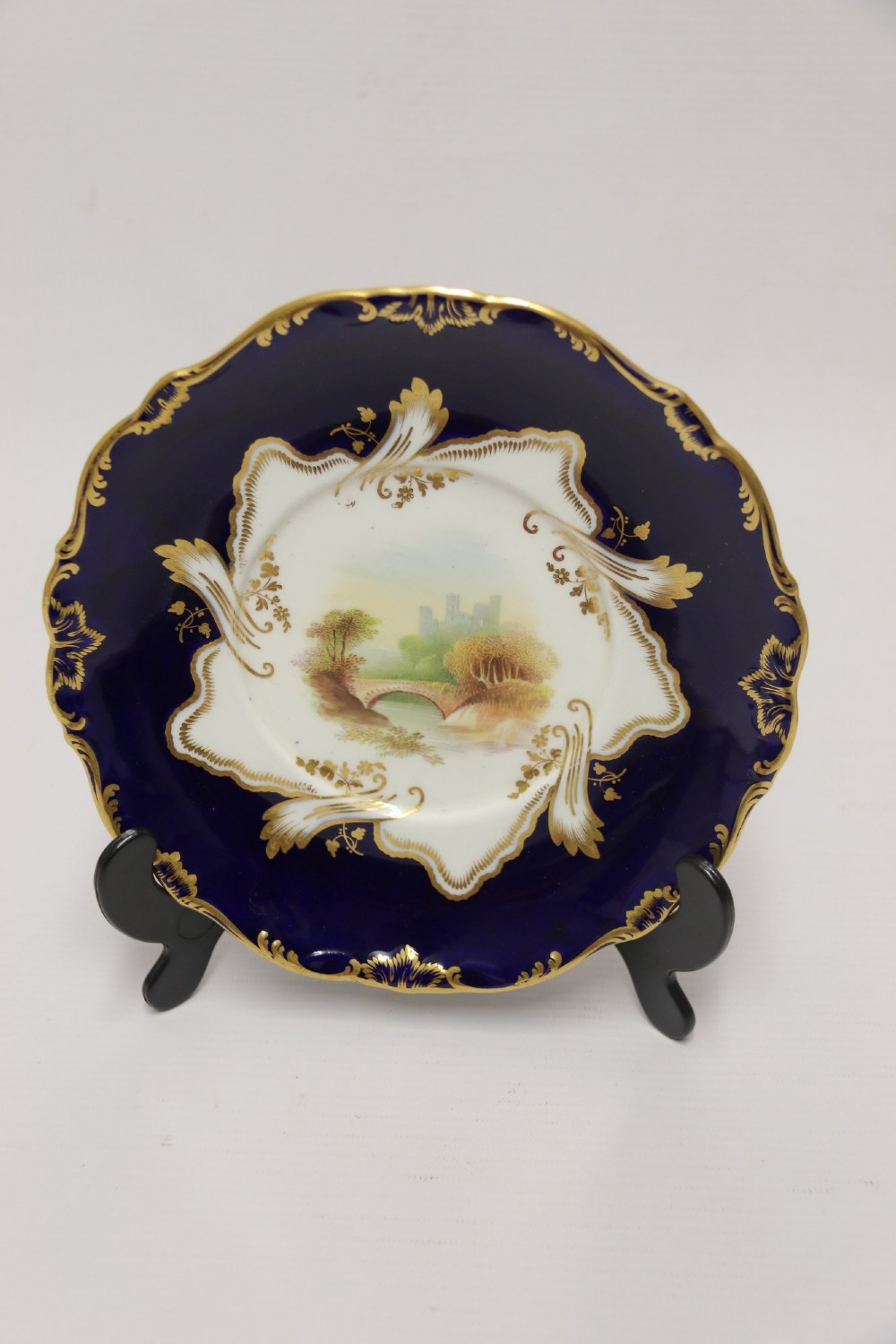 A 19th-century Mintons porcelain dessert cream tureen with lid and handles positioned alongside its matching stand, decorated with cobalt blue ground, gold gilding, and hand-painted landscape scenes. View V