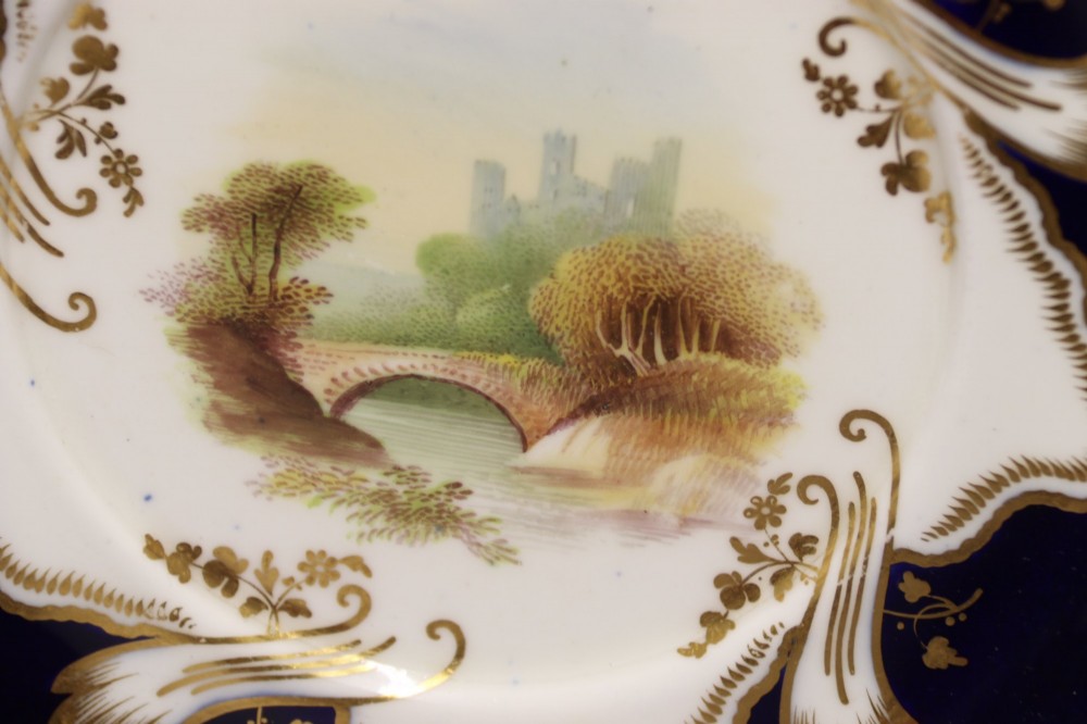 A 19th-century Mintons porcelain dessert cream tureen with lid and handles positioned alongside its matching stand, decorated with cobalt blue ground, gold gilding, and hand-painted landscape scenes. View VI