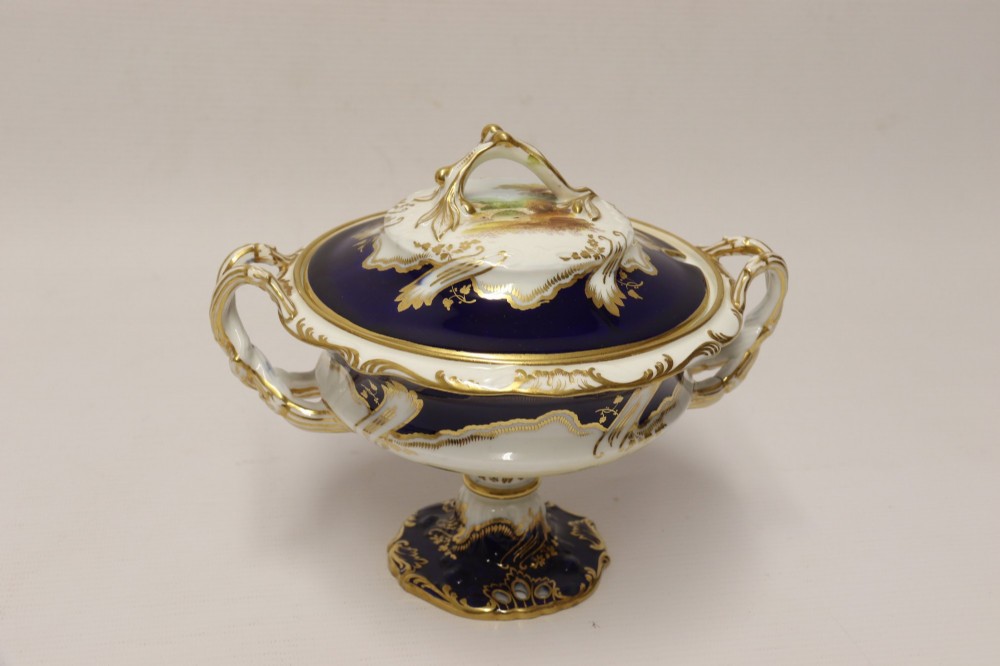 A 19th-century Mintons porcelain dessert cream tureen with lid and handles positioned alongside its matching stand, decorated with cobalt blue ground, gold gilding, and hand-painted landscape scenes. View VIII
