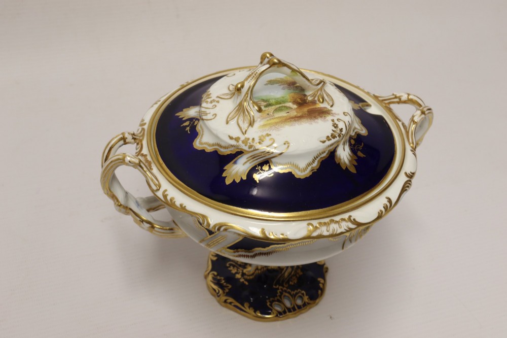 A 19th-century Mintons porcelain dessert cream tureen with lid and handles positioned alongside its matching stand, decorated with cobalt blue ground, gold gilding, and hand-painted landscape scenes. View IX