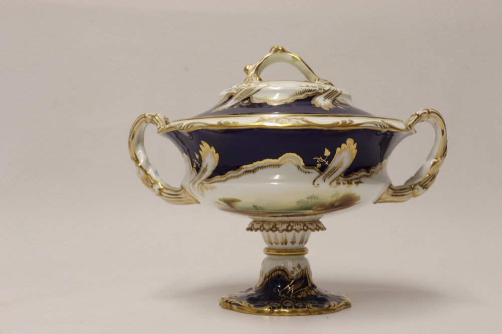 A 19th-century Mintons porcelain dessert cream tureen with lid and handles positioned alongside its matching stand, decorated with cobalt blue ground, gold gilding, and hand-painted landscape scenes. View X