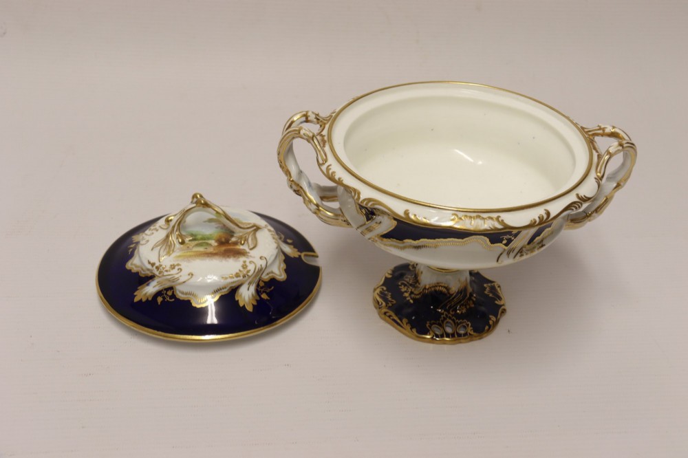 A 19th-century Mintons porcelain dessert cream tureen with lid and handles positioned alongside its matching stand, decorated with cobalt blue ground, gold gilding, and hand-painted landscape scenes. View XI