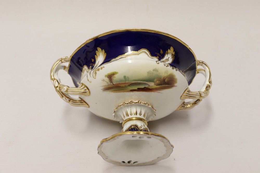 A 19th-century Mintons porcelain dessert cream tureen with lid and handles positioned alongside its matching stand, decorated with cobalt blue ground, gold gilding, and hand-painted landscape scenes. View XII
