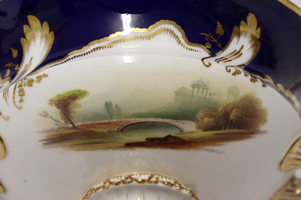 A 19th-century Mintons porcelain dessert cream tureen with lid and handles positioned alongside its matching stand, decorated with cobalt blue ground, gold gilding, and hand-painted landscape scenes. View XIII