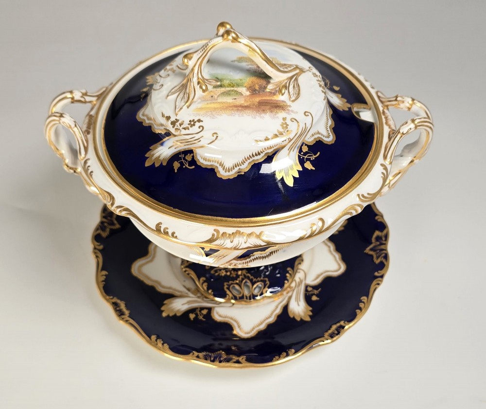 A 19th-century Mintons porcelain dessert cream tureen with lid and handles positioned alongside its matching stand, decorated with cobalt blue ground, gold gilding, and hand-painted landscape scenes. View XIV