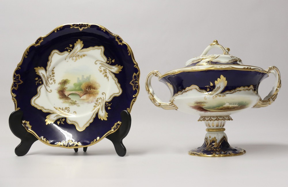 A 19th-century Mintons porcelain dessert cream tureen with lid and handles positioned alongside its matching stand, decorated with cobalt blue ground, gold gilding, and hand-painted landscape scenes. View XV