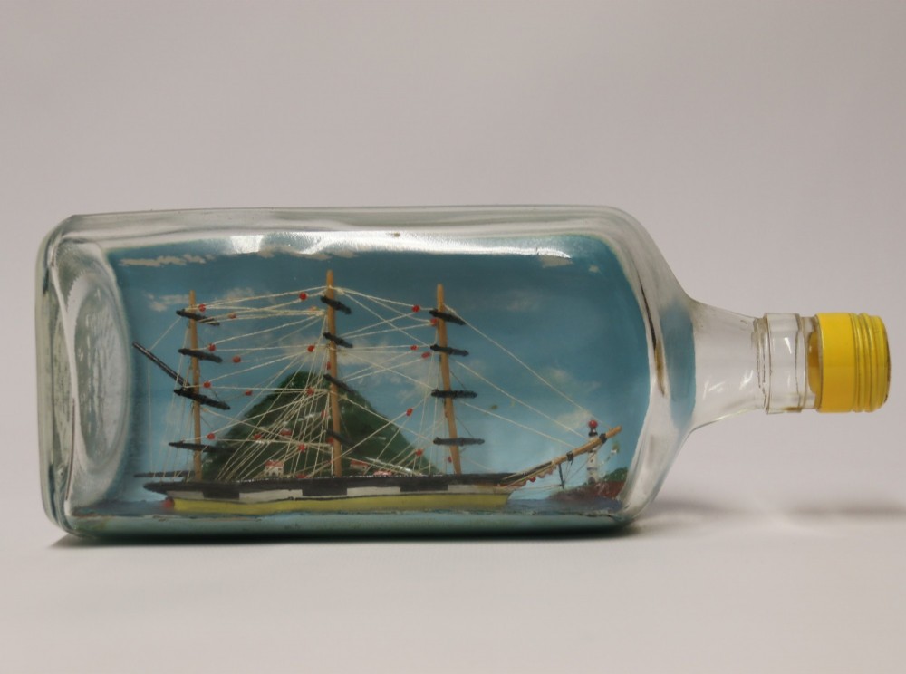 A miniature three-masted wooden sailing vessel set inside a horizontal clear glass bottle against a blue painted backdrop.