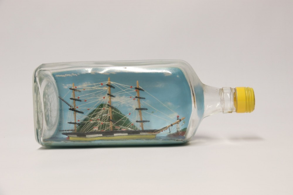 A miniature three-masted wooden sailing vessel set inside a horizontal clear glass bottle against a blue painted backdrop. View II