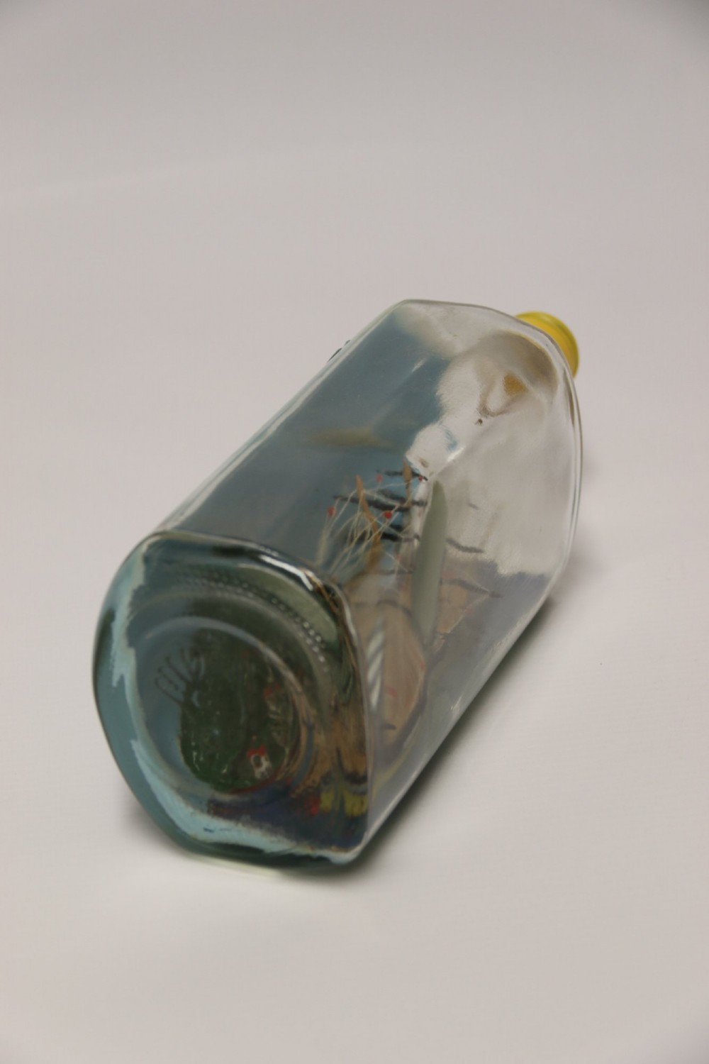 A miniature three-masted wooden sailing vessel set inside a horizontal clear glass bottle against a blue painted backdrop. View VI
