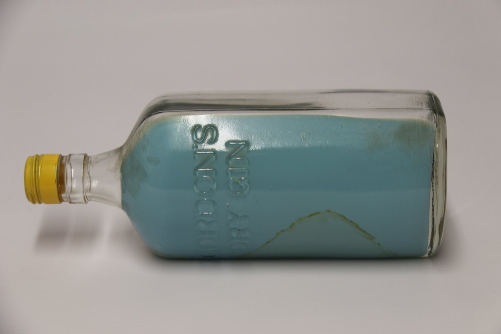 A miniature three-masted wooden sailing vessel set inside a horizontal clear glass bottle against a blue painted backdrop. View VII