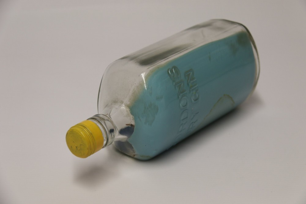 A miniature three-masted wooden sailing vessel set inside a horizontal clear glass bottle against a blue painted backdrop. View VIII