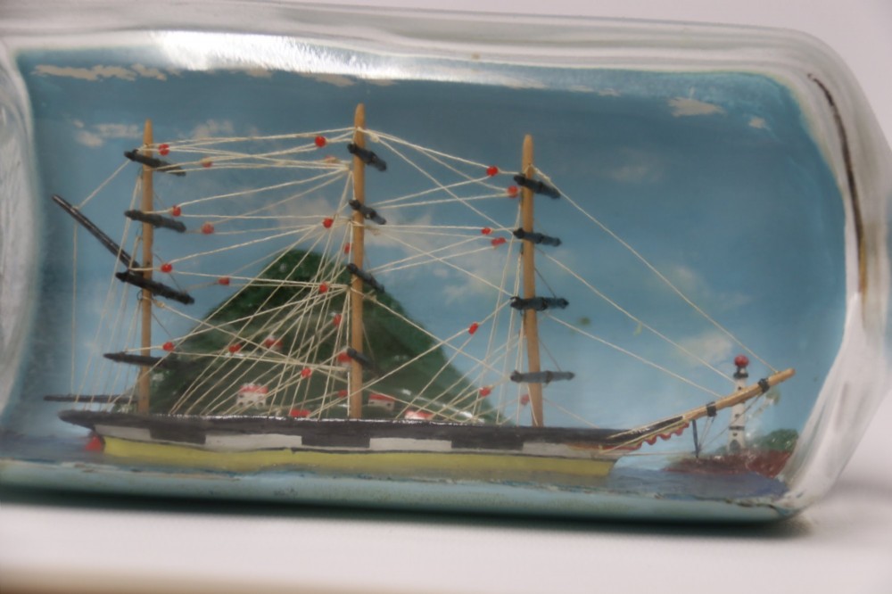 A miniature three-masted wooden sailing vessel set inside a horizontal clear glass bottle against a blue painted backdrop. View IX