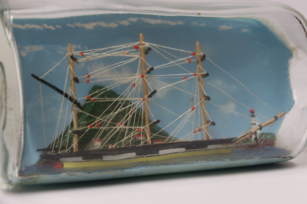 A miniature three-masted wooden sailing vessel set inside a horizontal clear glass bottle against a blue painted backdrop. View X