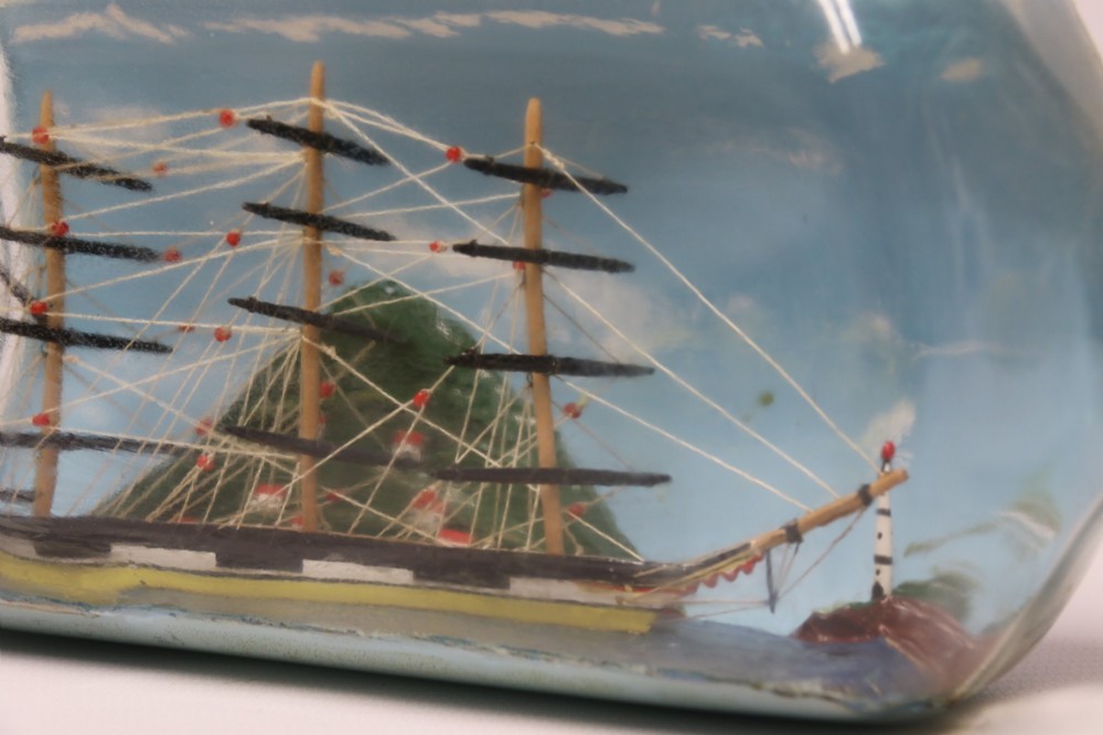A miniature three-masted wooden sailing vessel set inside a horizontal clear glass bottle against a blue painted backdrop. View XI