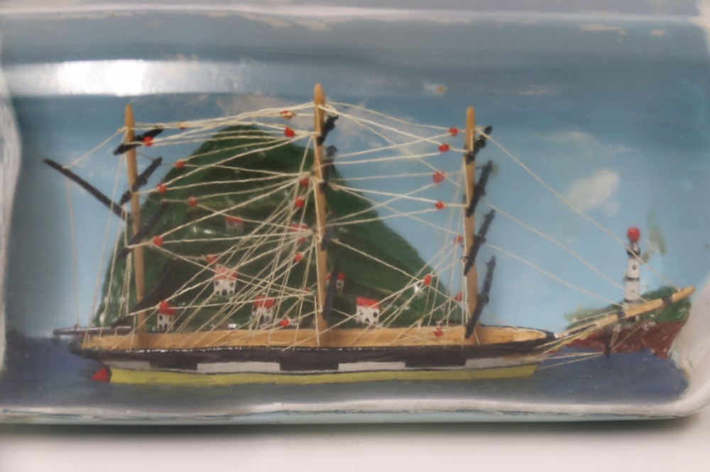 A miniature three-masted wooden sailing vessel set inside a horizontal clear glass bottle against a blue painted backdrop. View XII
