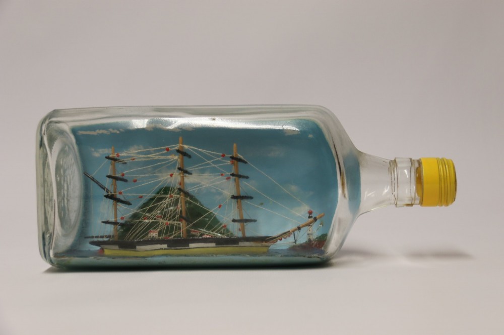 A miniature three-masted wooden sailing vessel set inside a horizontal clear glass bottle against a blue painted backdrop. View XIII