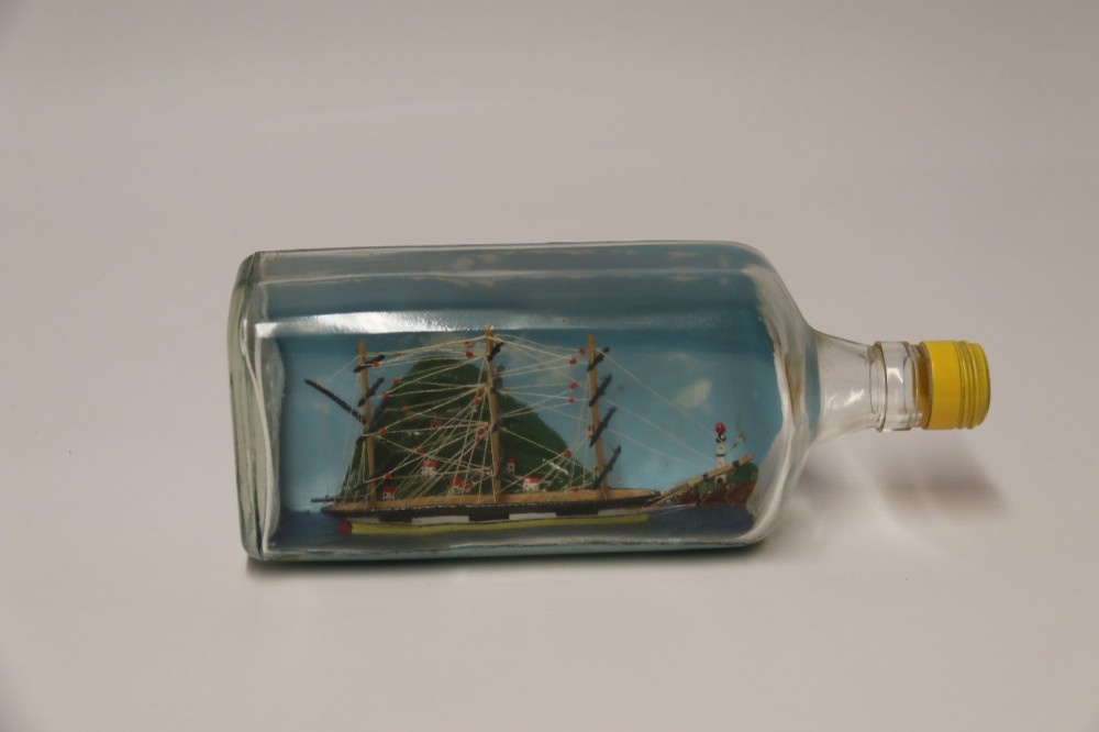 A miniature three-masted wooden sailing vessel set inside a horizontal clear glass bottle against a blue painted backdrop. View XIV