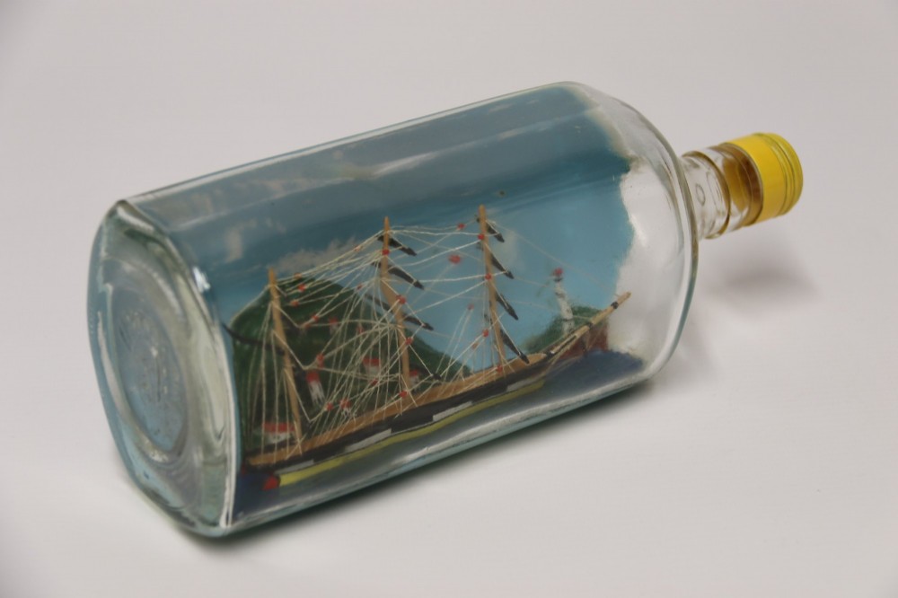 A miniature three-masted wooden sailing vessel set inside a horizontal clear glass bottle against a blue painted backdrop. View XV