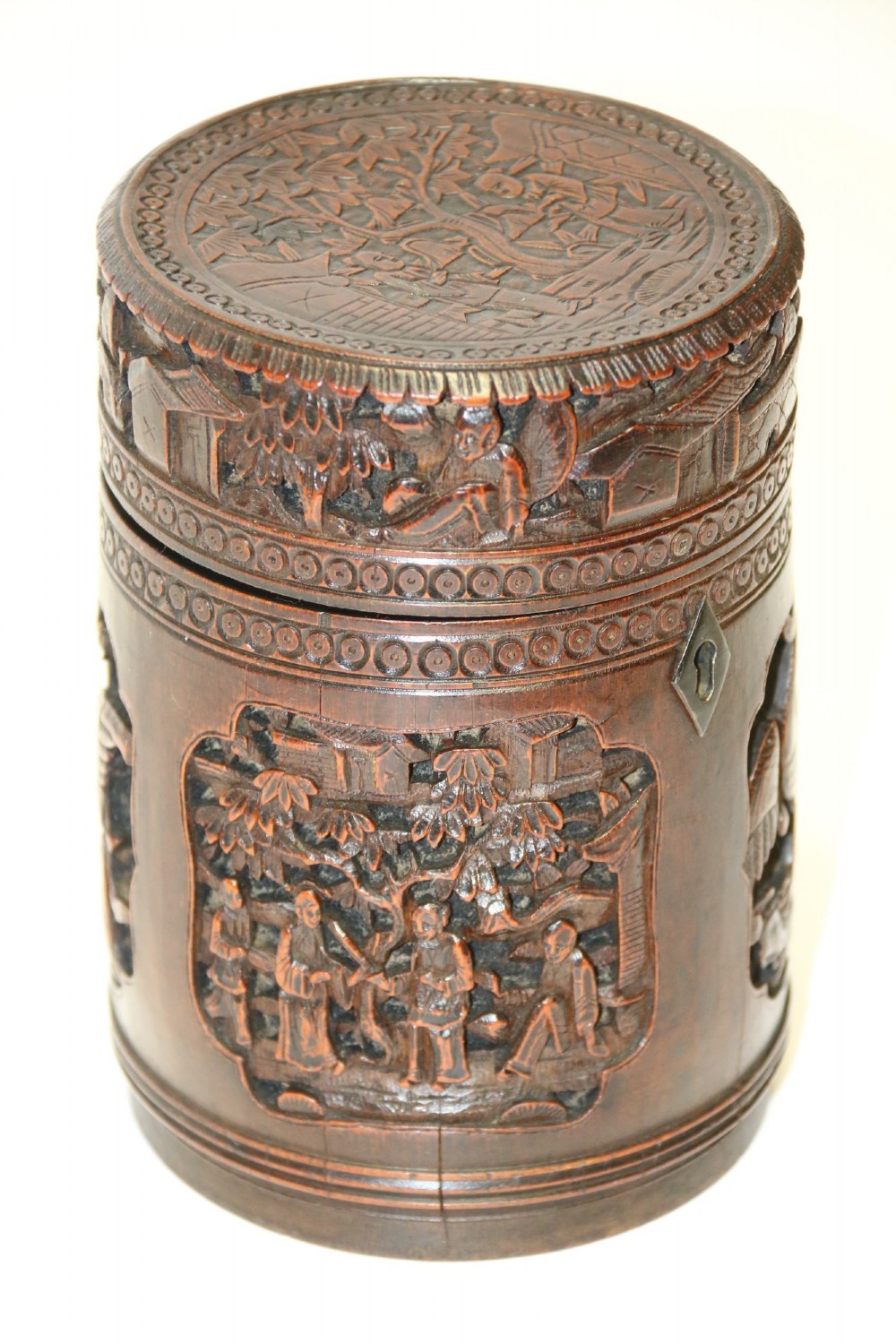 A cylindrical dark brown bamboo tea caddy featuring intricate high-relief carvings of Chinese figures, trees, and architecture with a metal escutcheon.