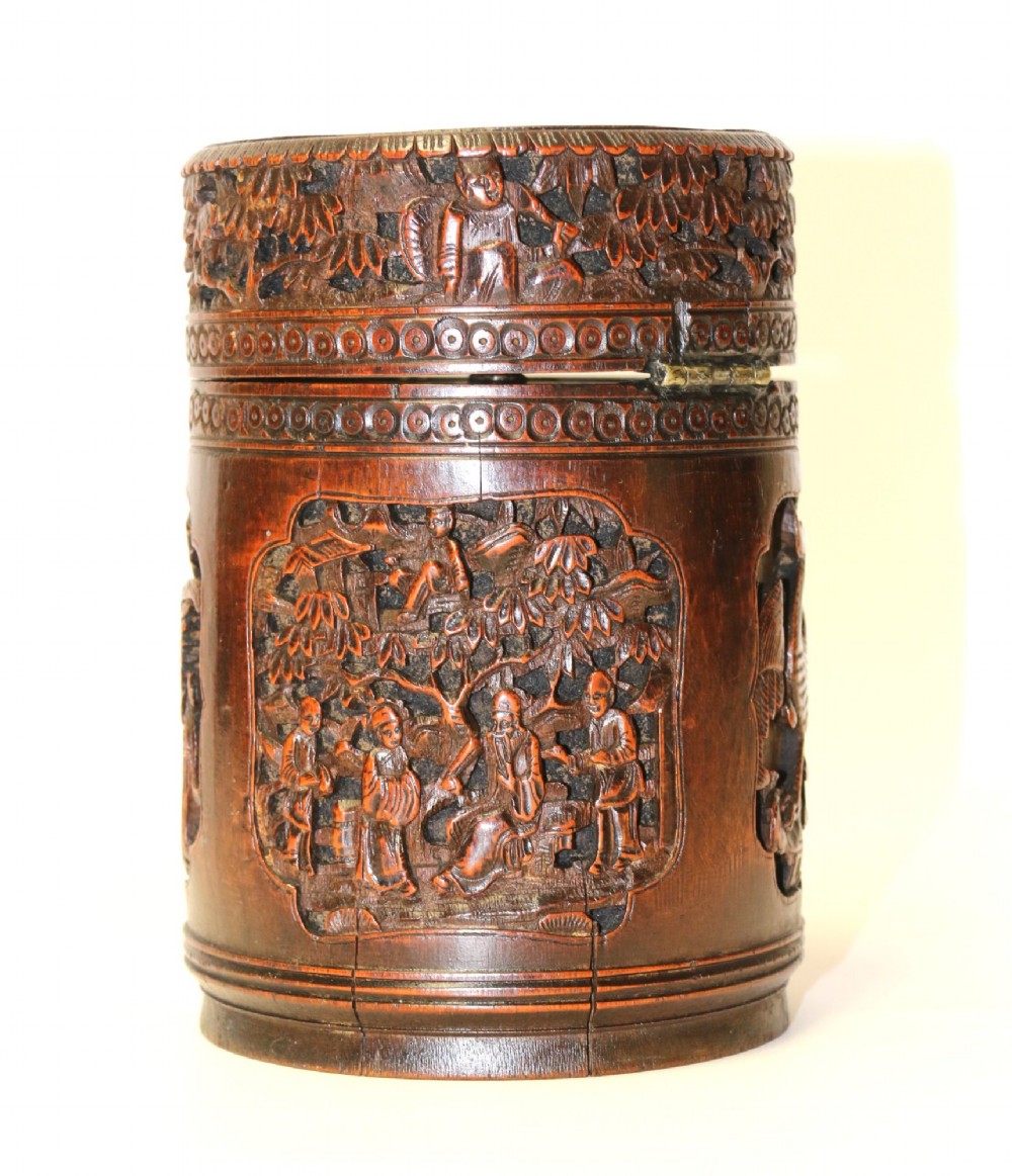 A cylindrical dark brown bamboo tea caddy featuring intricate high-relief carvings of Chinese figures, trees, and architecture with a metal escutcheon. View II