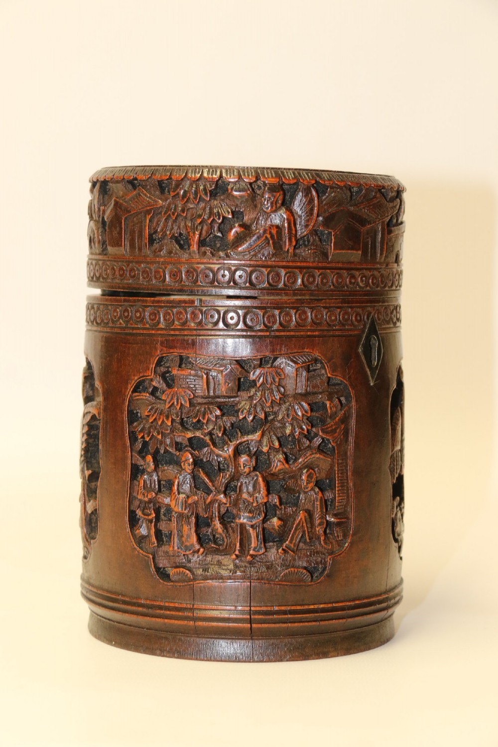 A cylindrical dark brown bamboo tea caddy featuring intricate high-relief carvings of Chinese figures, trees, and architecture with a metal escutcheon. View III