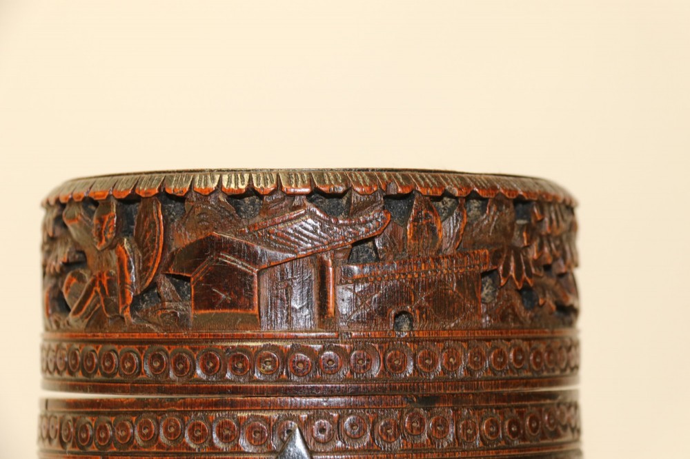 A cylindrical dark brown bamboo tea caddy featuring intricate high-relief carvings of Chinese figures, trees, and architecture with a metal escutcheon. View IV