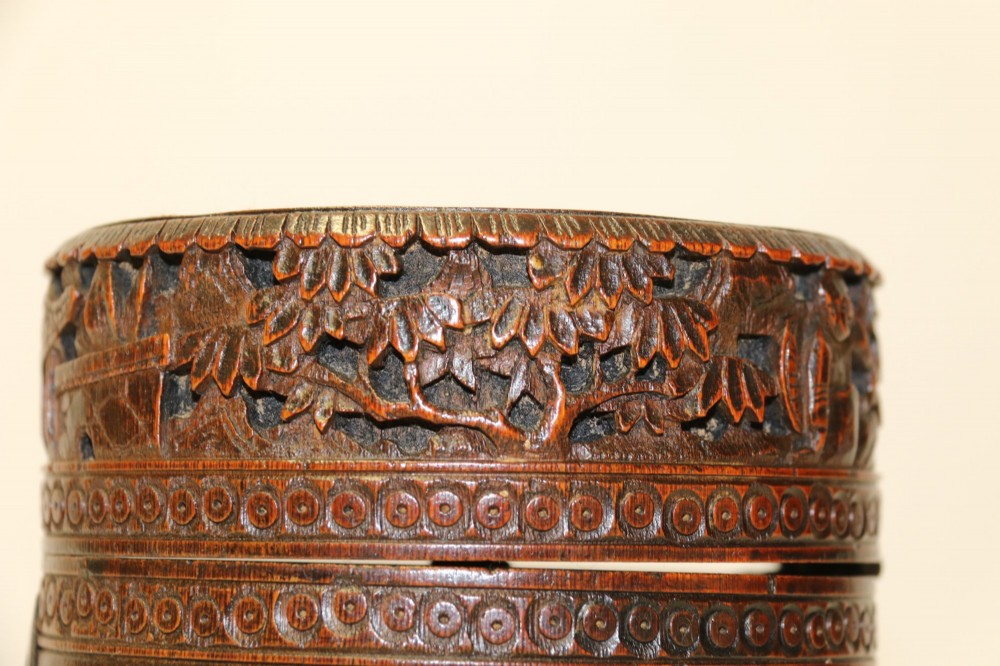 A cylindrical dark brown bamboo tea caddy featuring intricate high-relief carvings of Chinese figures, trees, and architecture with a metal escutcheon. View V