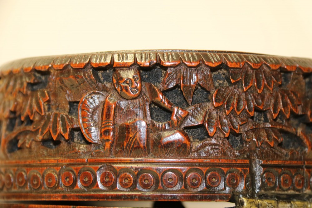 A cylindrical dark brown bamboo tea caddy featuring intricate high-relief carvings of Chinese figures, trees, and architecture with a metal escutcheon. View VI