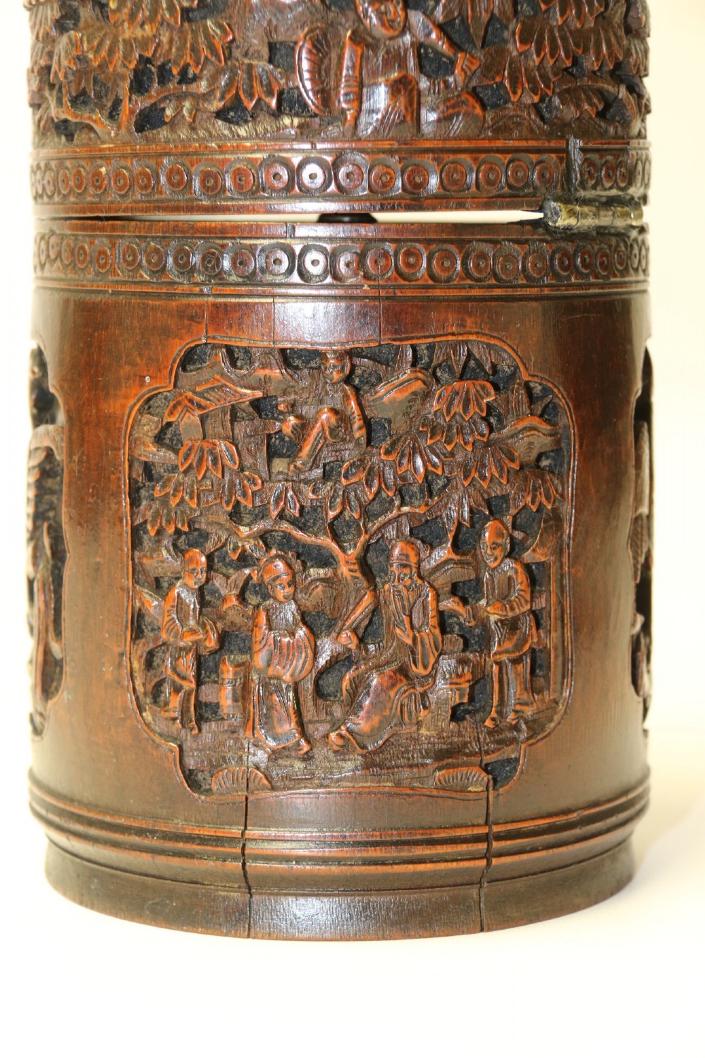 A cylindrical dark brown bamboo tea caddy featuring intricate high-relief carvings of Chinese figures, trees, and architecture with a metal escutcheon. View VII