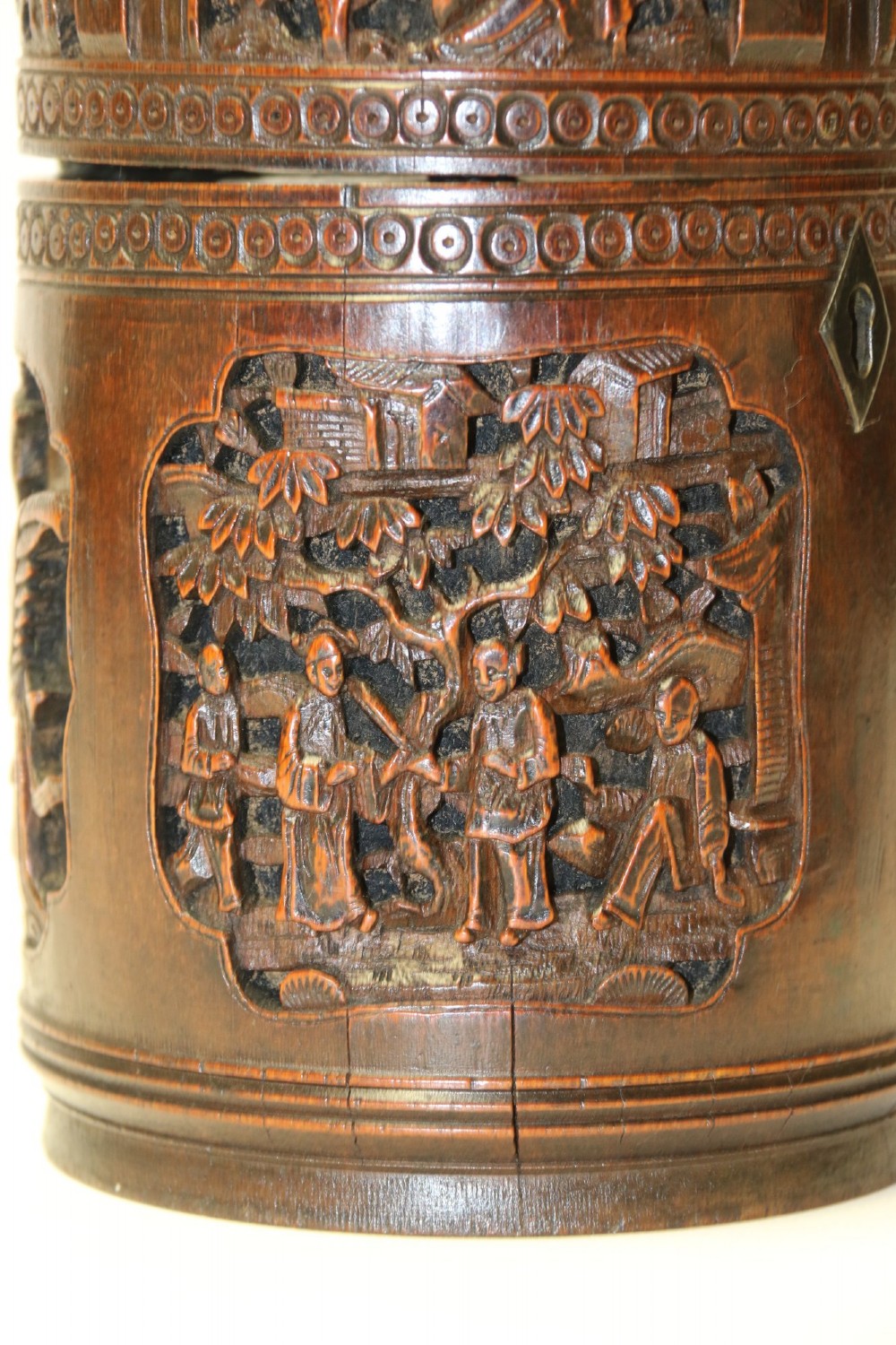 A cylindrical dark brown bamboo tea caddy featuring intricate high-relief carvings of Chinese figures, trees, and architecture with a metal escutcheon. View VIII