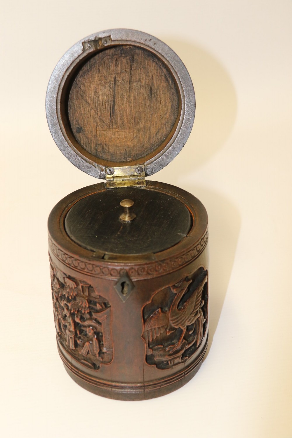 A cylindrical dark brown bamboo tea caddy featuring intricate high-relief carvings of Chinese figures, trees, and architecture with a metal escutcheon. View X