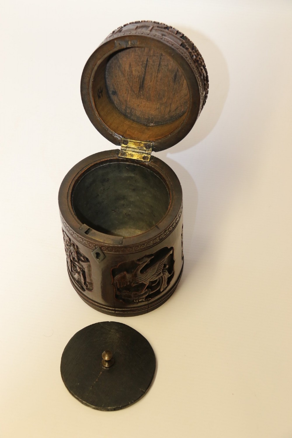 A cylindrical dark brown bamboo tea caddy featuring intricate high-relief carvings of Chinese figures, trees, and architecture with a metal escutcheon. View XI
