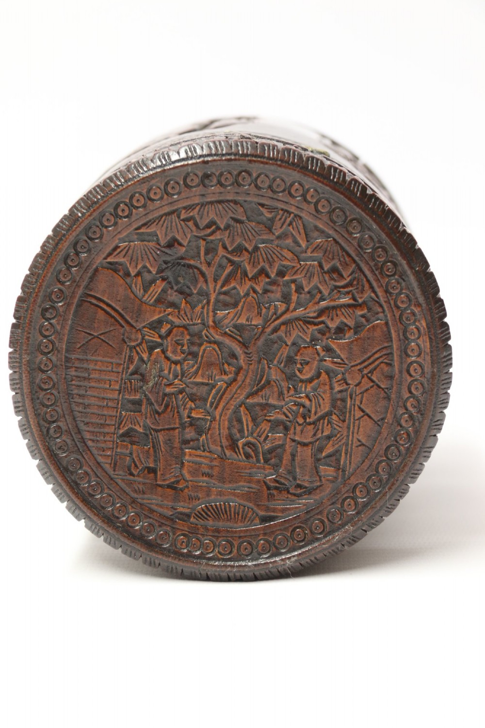 A cylindrical dark brown bamboo tea caddy featuring intricate high-relief carvings of Chinese figures, trees, and architecture with a metal escutcheon. View XII
