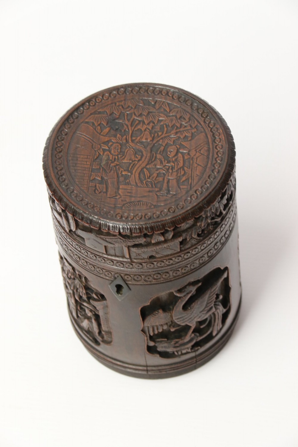 A cylindrical dark brown bamboo tea caddy featuring intricate high-relief carvings of Chinese figures, trees, and architecture with a metal escutcheon. View XIII