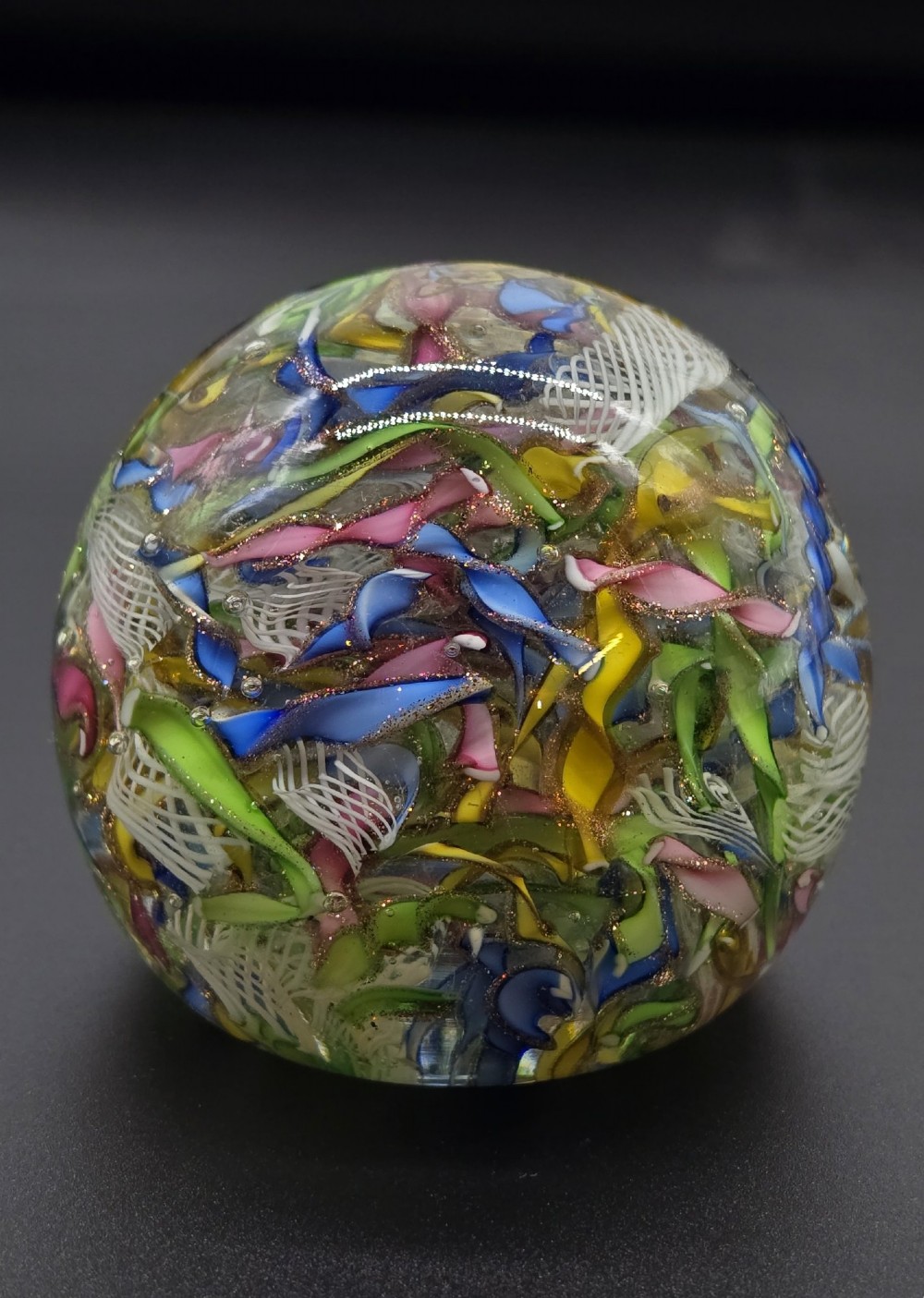 A clear spherical glass paperweight filled with an intricate mix of colourful cane fragments, white filigree ribbons, and glittering gold aventurine flecks. View II